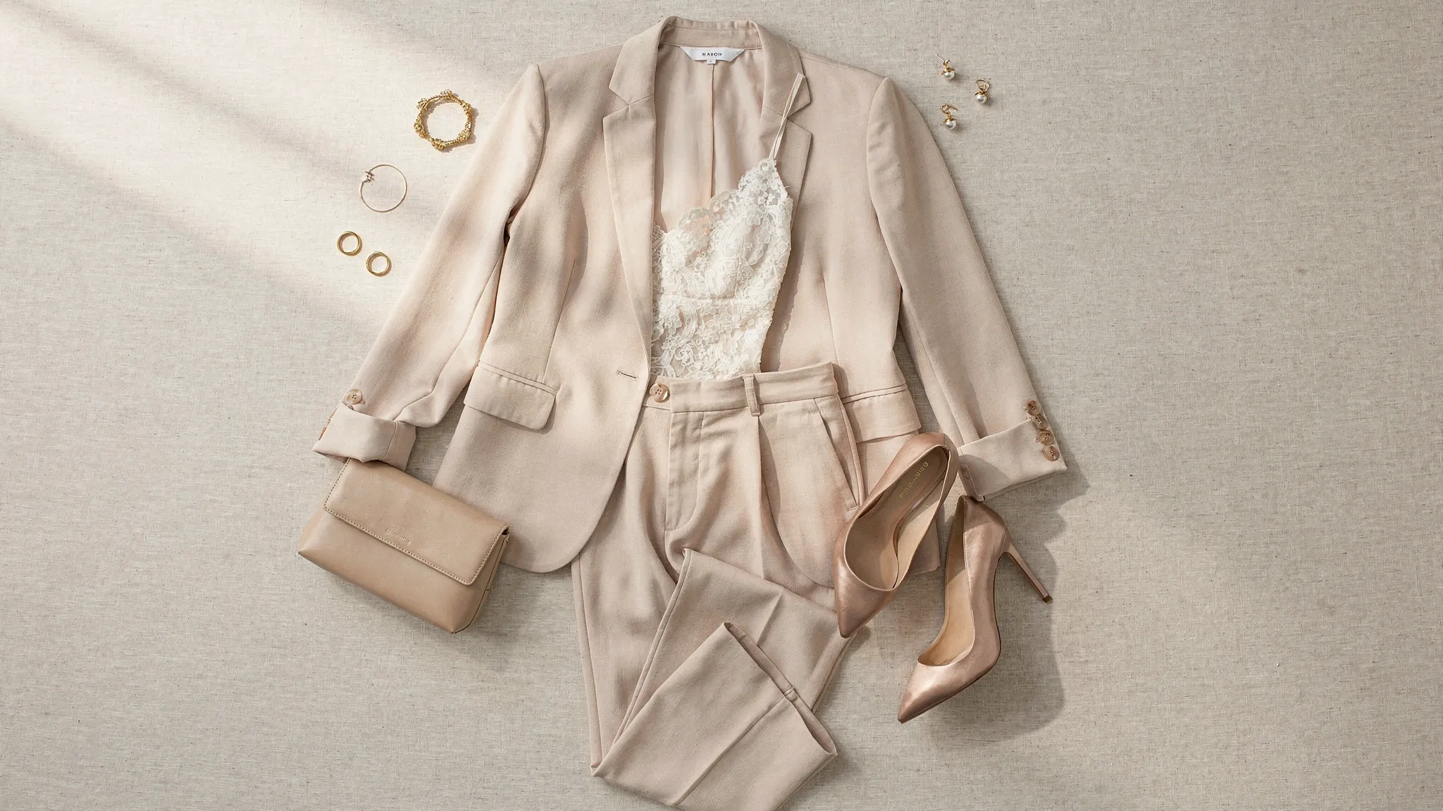 Een flatlay met een trouwproof broekpak outfit: blazer en pantalon in zachte champagne tint, kanten top, hakken, clutch en subtiele sieraden op een neutrale achtergrond.